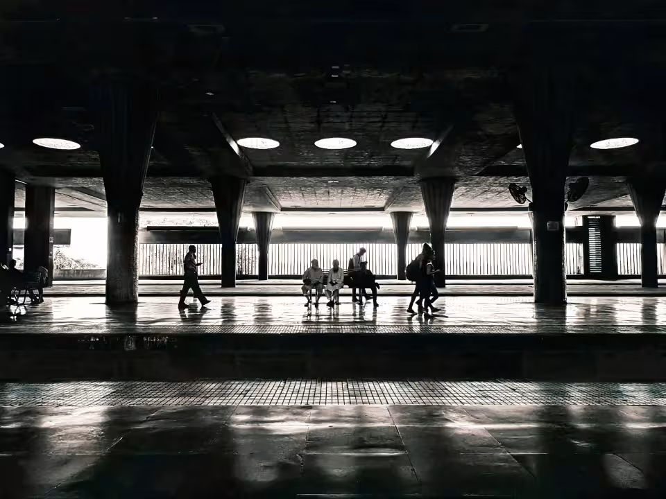 Silhuetas de pessoas esperando em uma estação de trem pouco iluminada, com janelas claras.
